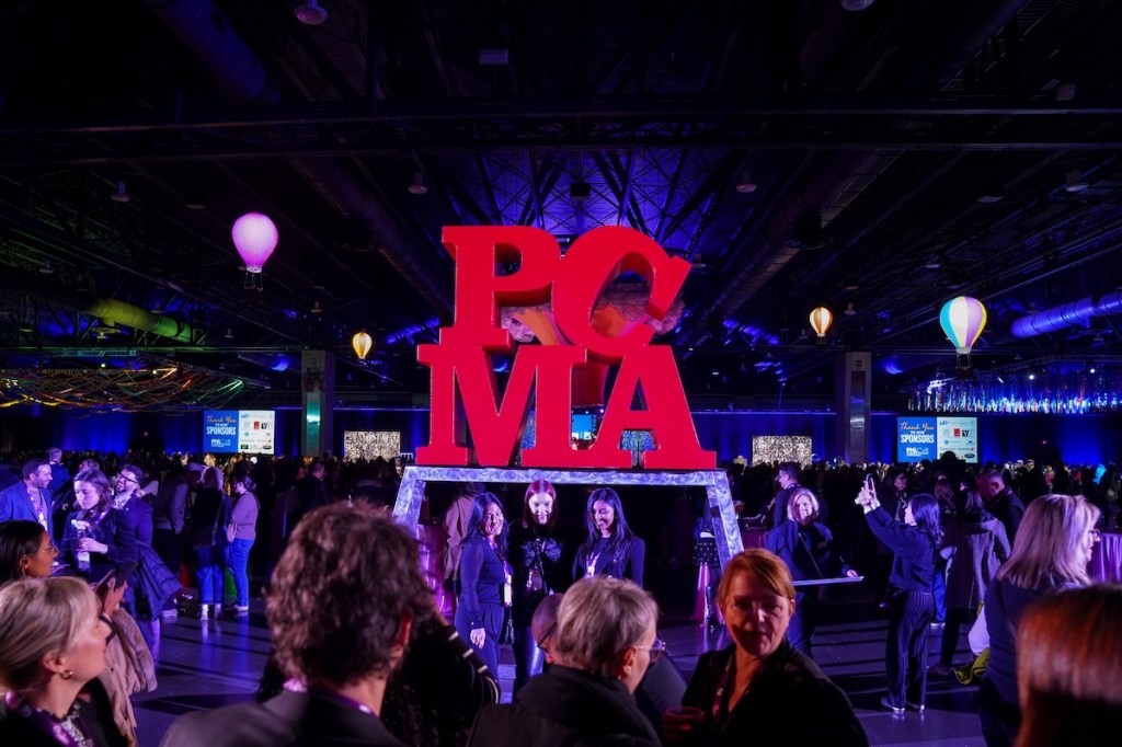 People taking photos in front of a temporary PCMA statue in bright red in the style of LOVE, by Robert Indiana, inside a convention center hall